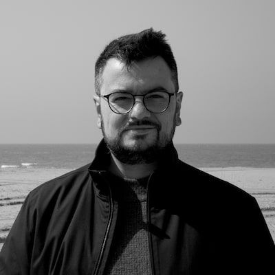 Front-facing black and white medium close-up photo of Nicolas Magand, wearing glasses, with a beach in the background.